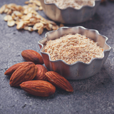 Small stainless steel flan dish of Almond Flour sitting on concrete bench-top next to five whole raw almonds.