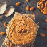 Almond Butter in round glass bowl with scattered whole almonds and silver spoons. Sitting on rustic black timber table.