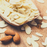 Timber serving spoon full of Blanched Almond Flakes sitting next to three raw whole almonds.