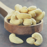 Raw Cashews Nuts spilling over rustic timber serving spoon, on dark grey background. 