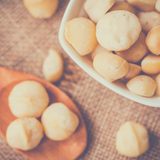 Macadamia Nuts Raw in white bowl, sitting on top of hessian background with timber serving spoon holding three whole raw macadamias.