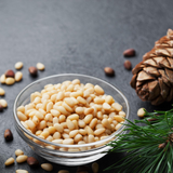 Small glass bowl of Pine Nuts on dark background. Pine Nuts scattered around the bowl and pine cone in background.