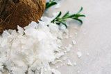 Coconut Flakes piled up next to whole coconut, with sprigs of green leaves on light grey background.