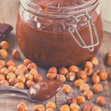 Glass jar of Chocolate Hazelnut Butter sitting next to spoon and loose whole hazelnuts.
