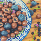 Milk Chocolate Covered Blueberries mixed with dried blueberries and purple flowers, sitting in blue patterned bowl. Timber background.