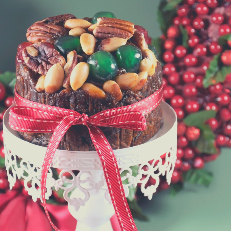 Small Chistmas Cake on white stand, topped with pecans, brazil nuts and Glace Cherries Green.