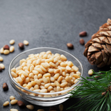 Small glass bowl of Pine Nuts on dark background. Pine Nuts scattered around the bowl and pine cone in background. 