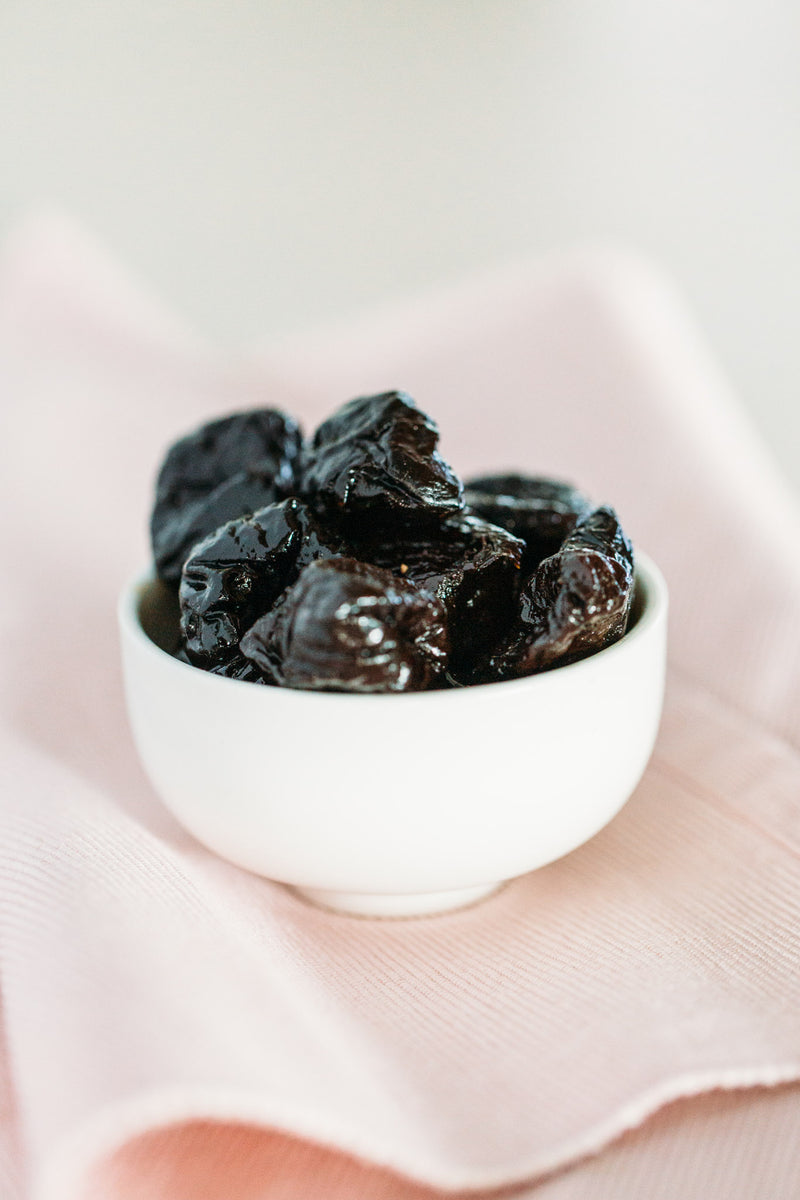 Prunes in a small ceramic bowl on soft pink cloth.