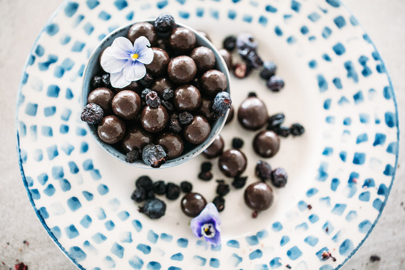 Small bowl of Dark Chocolate Blueberries stacked on white and blue plate. Decorated with purple edible flowers. 