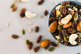 Close up of Healthy Trail Mix in small blue bowl, surrounded by scattered hikers mix on concrete background.