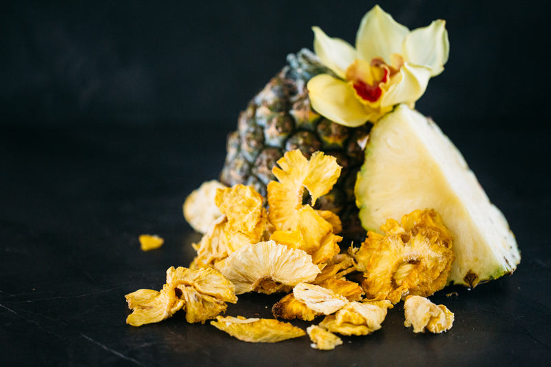 Dried Pineapple scattered around whole fresh pineapple, on black stone benchtop.