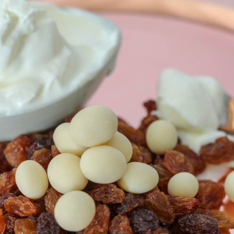 Close Up of Yoghurt Coated Sultanas sitting on top of sultanas with bowl of yoghurt.